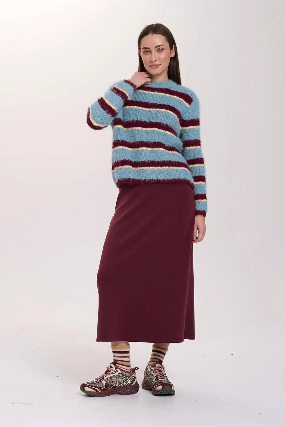 A model stands against a plain light background wearing a fuzzy striped sweater with light blue, beige, and burgundy stripes, paired with a solid burgundy midi skirt and patterned sneakers.