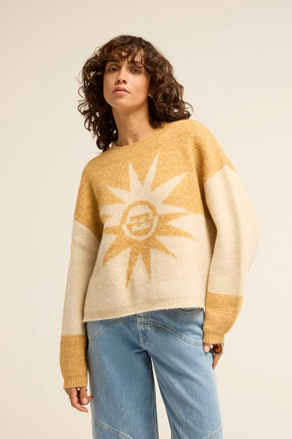 A woman wearing a chunky knit sweater with a large sun graphic on the front, paired with light blue jeans, against a plain beige studio background.
