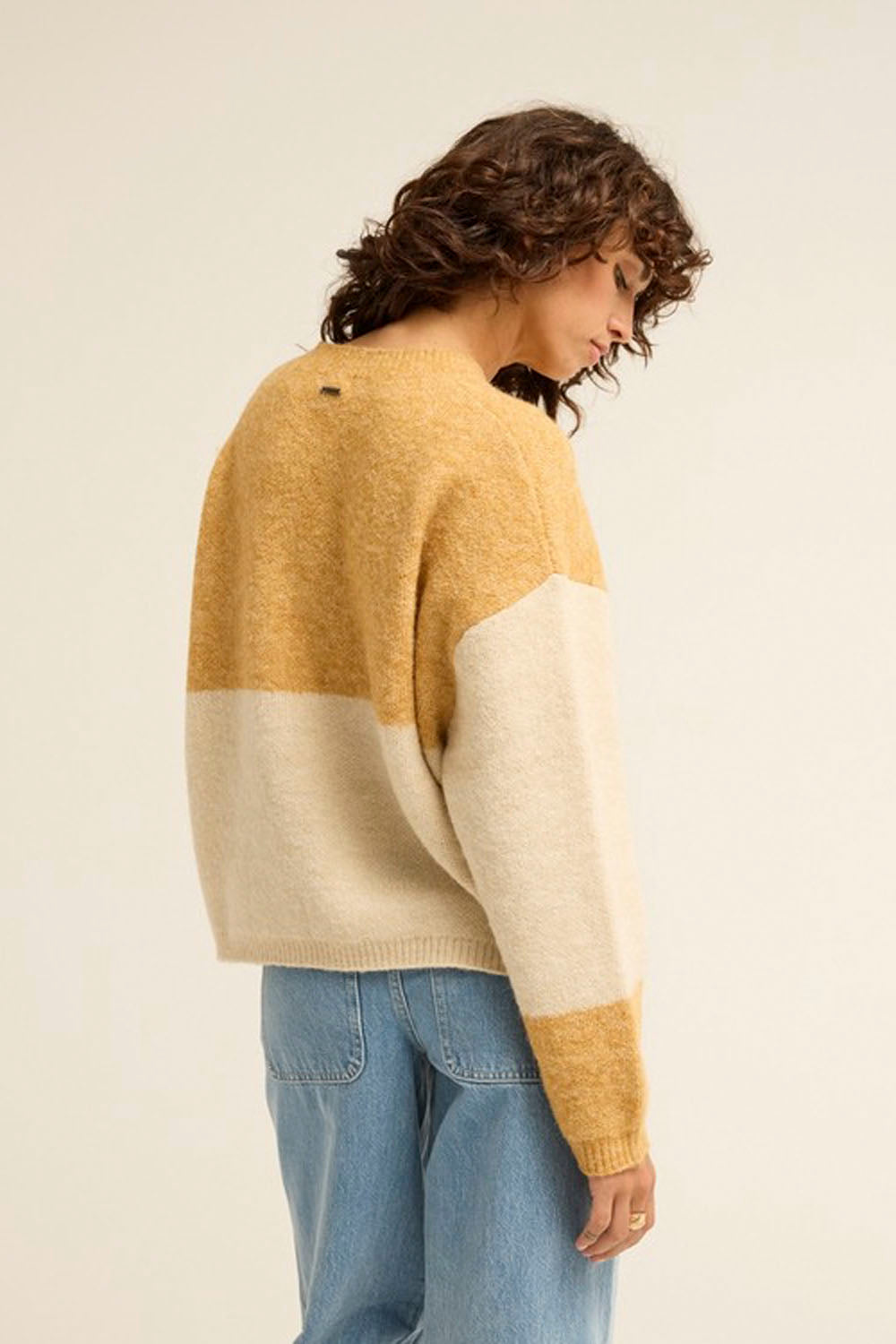 A woman wearing a chunky knit color-block sweater in mustard yellow and cream, paired with light blue jeans, photographed from a side-back angle against a plain off-white studio background.