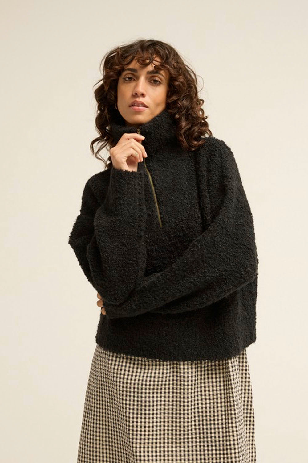 A woman wearing a black fuzzy turtleneck sweater and a black-and-white houndstooth skirt, standing against a plain light beige studio background.