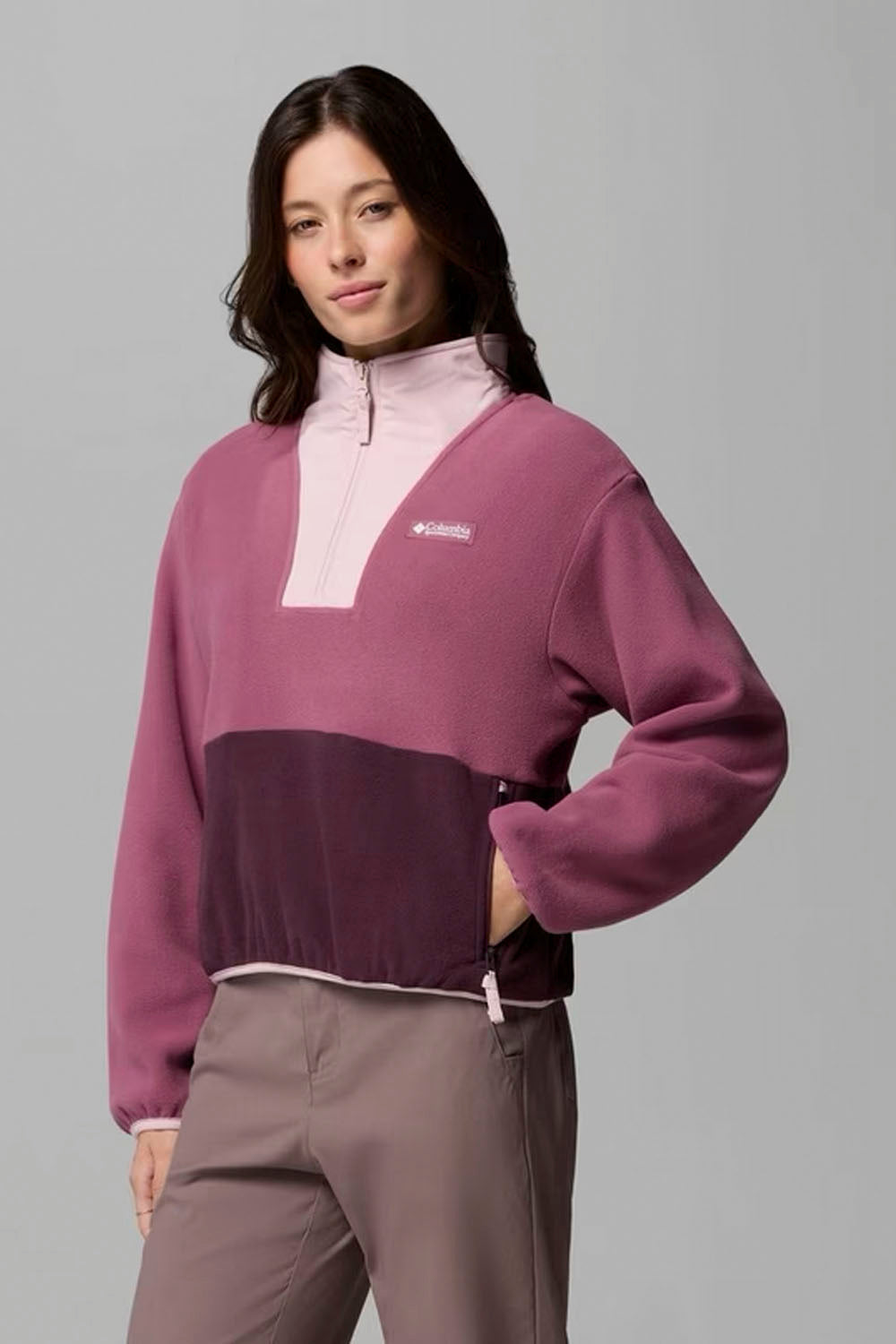 A woman modeling a color-block fleece pullover sweater with a light pink upper section, a dusty purple body, and a dark purple lower panel, paired with taupe pants against a solid gray studio background.