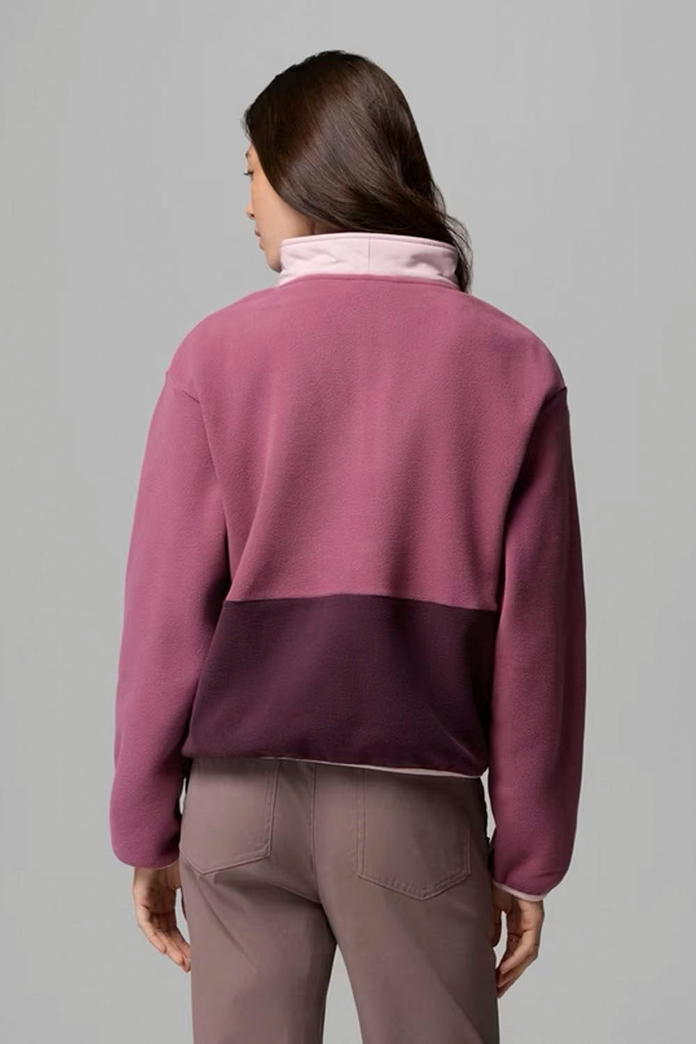 A woman shown from the back wearing a color-block fleece pullover with a dusty rose upper section and a dark plum lower section, paired with taupe pants, against a solid gray studio background.