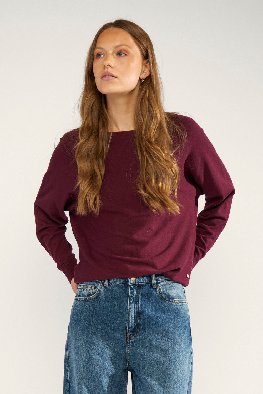 A woman wearing a loose-fitting, long-sleeve crewneck sweatshirt in deep burgundy, paired with blue jeans, photographed against a neutral light gray studio background.