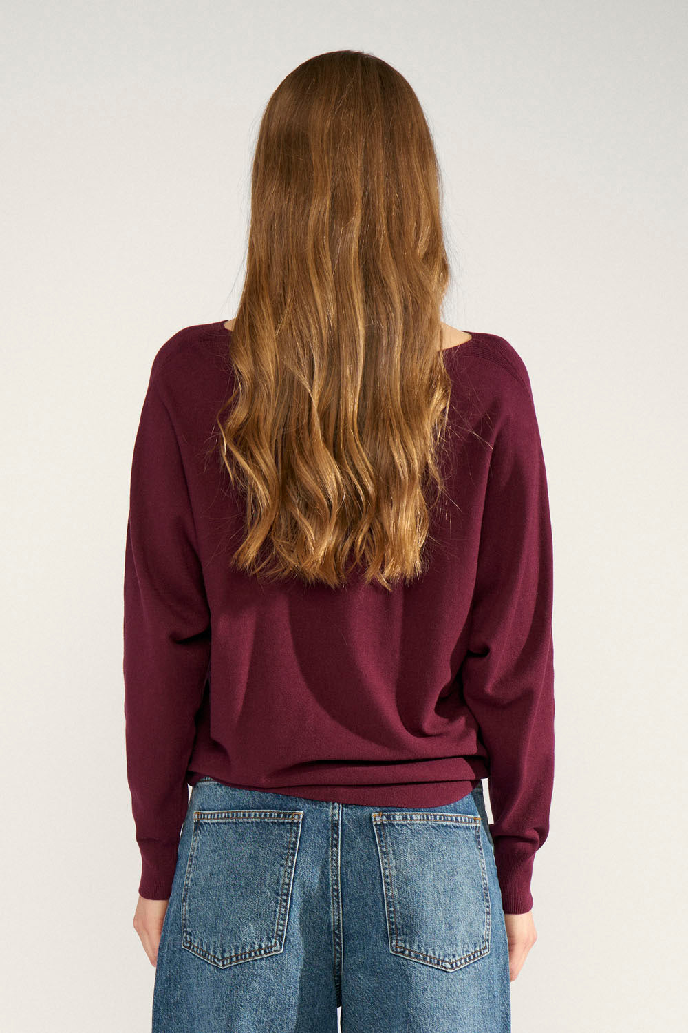 A woman viewed from behind wearing a long-sleeve maroon knit top and blue jeans against a plain light gray studio background.