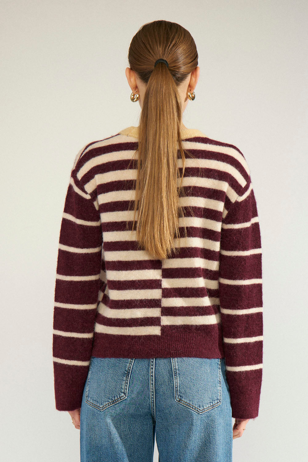 A woman viewed from behind wearing a maroon and cream striped knit sweater and blue jeans against a plain light gray background.