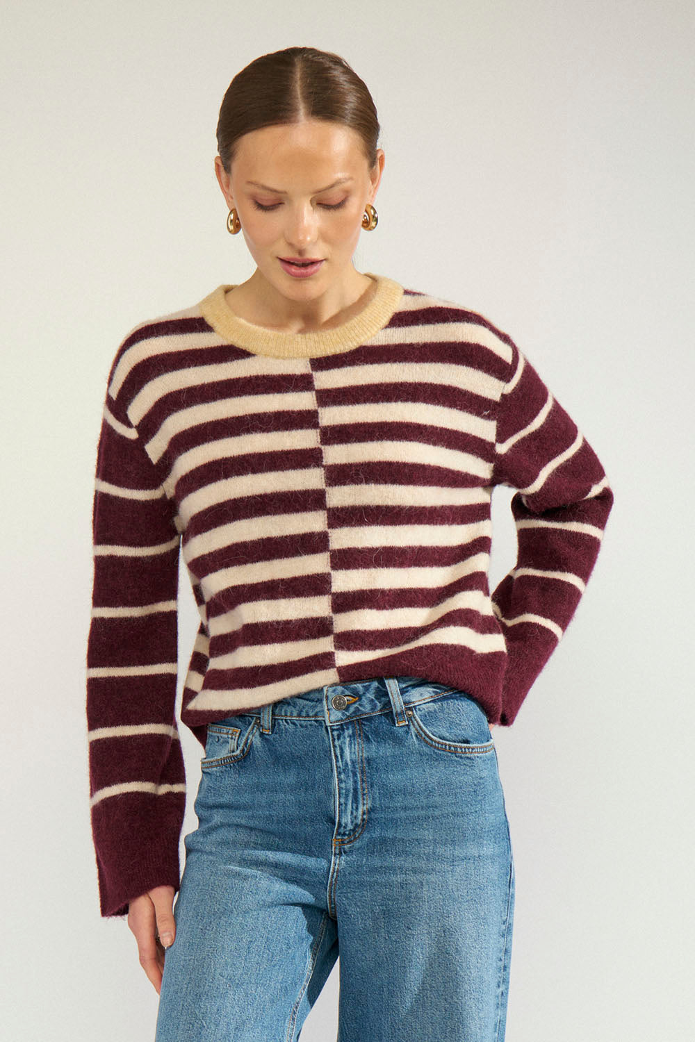 A woman wearing a fuzzy knit sweater with horizontal burgundy and cream stripes, paired with blue jeans, photographed against a light gray studio background.
