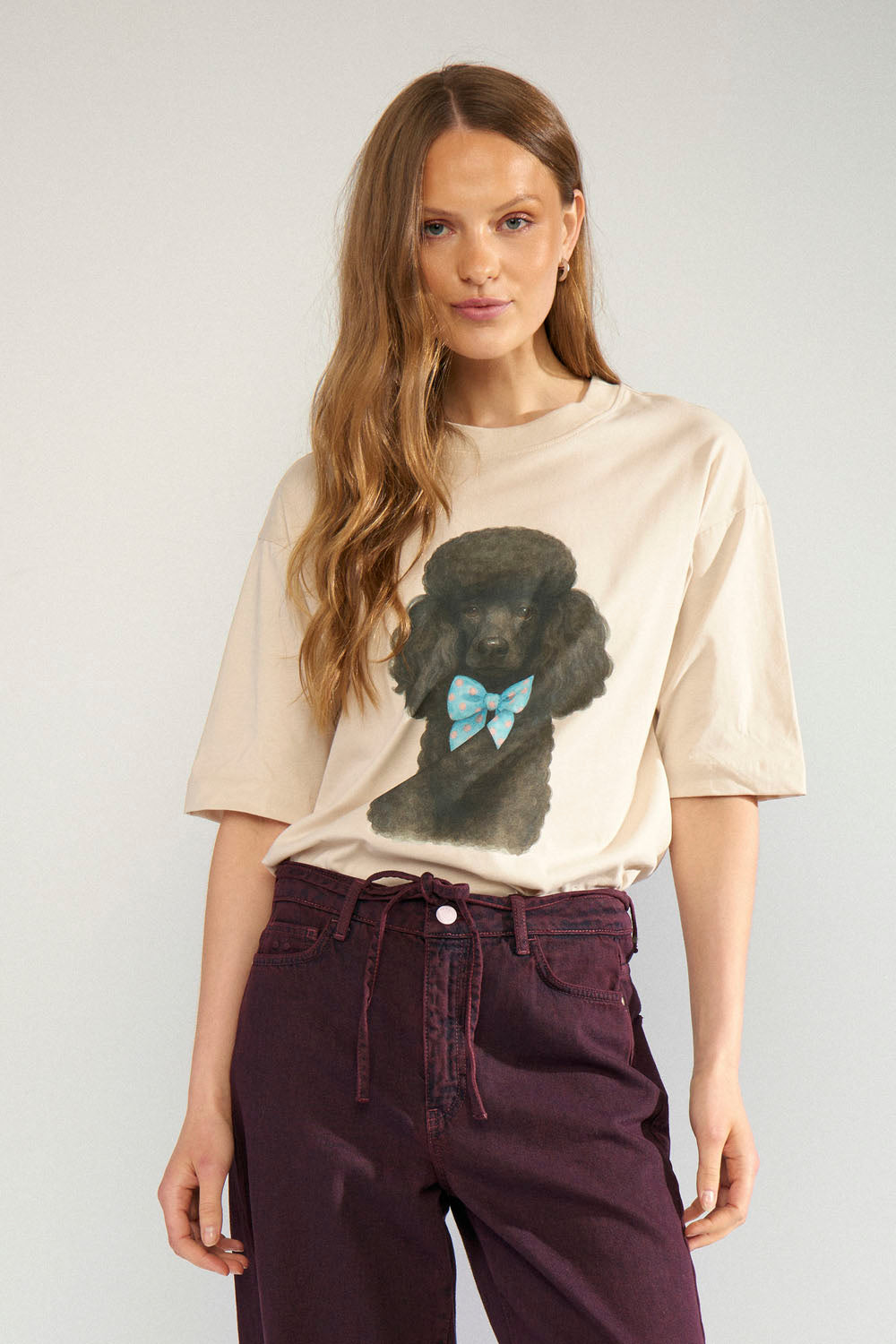 A woman modeling a beige graphic t-shirt featuring a black poodle wearing a light blue bow, paired with dark purple pants, against a light gray studio background.