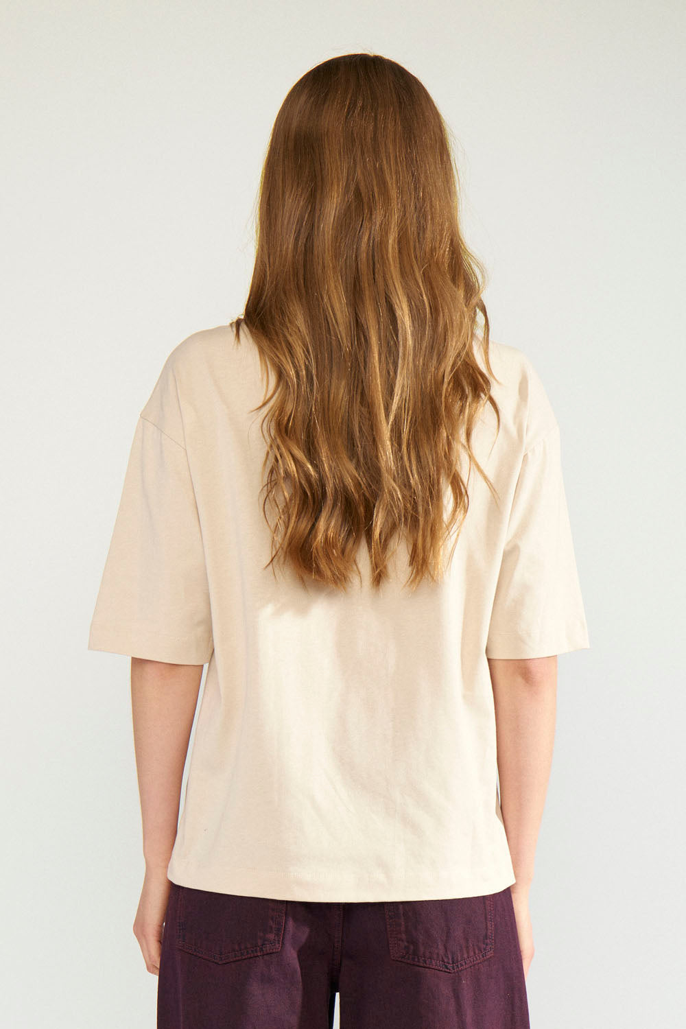 A woman viewed from behind wearing a cream-colored short-sleeve t-shirt and dark purple pants, set against a plain light gray studio background.