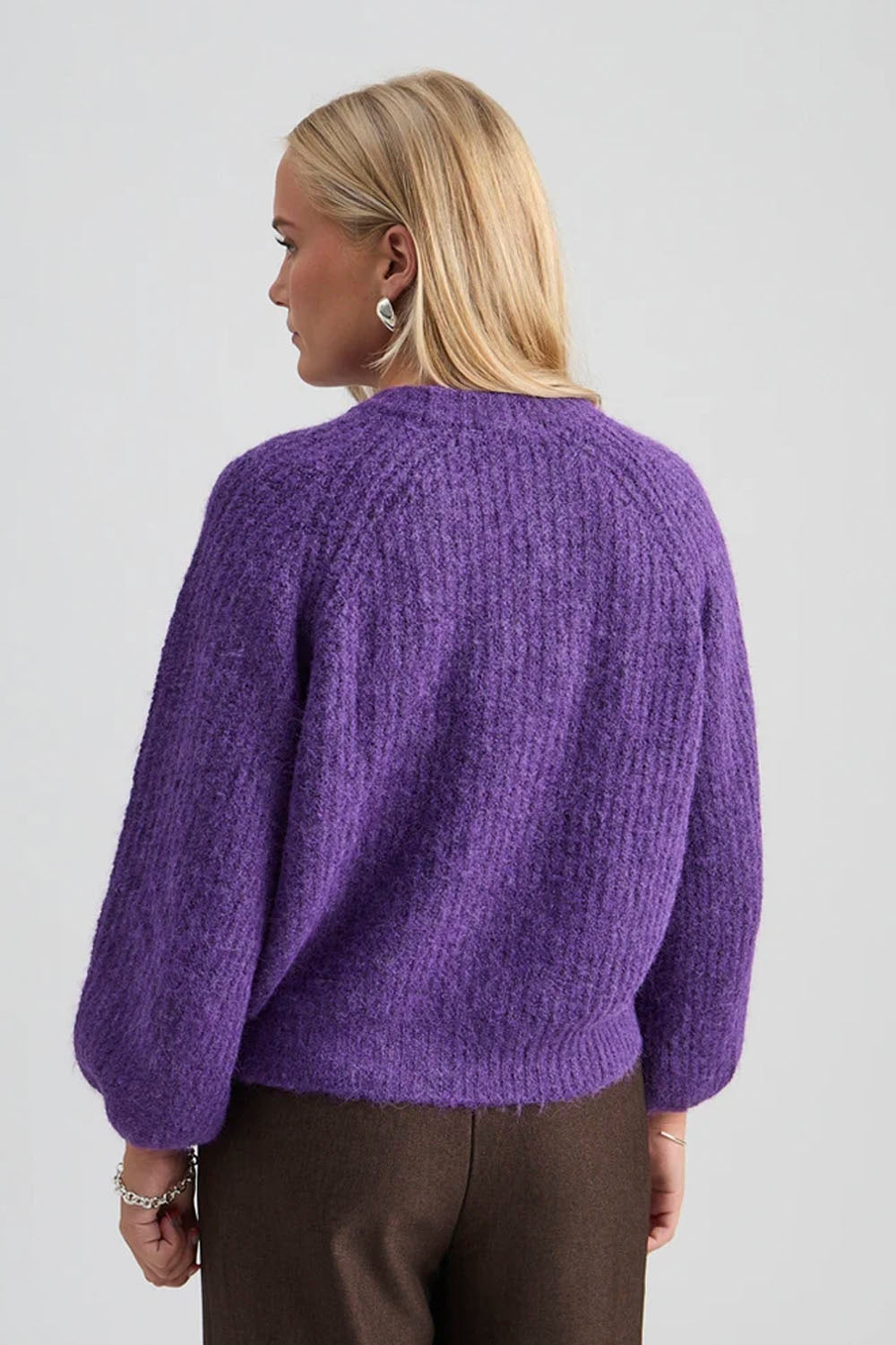 A woman shown from the back wearing a chunky, fuzzy purple knit sweater and brown trousers against a light gray studio background.