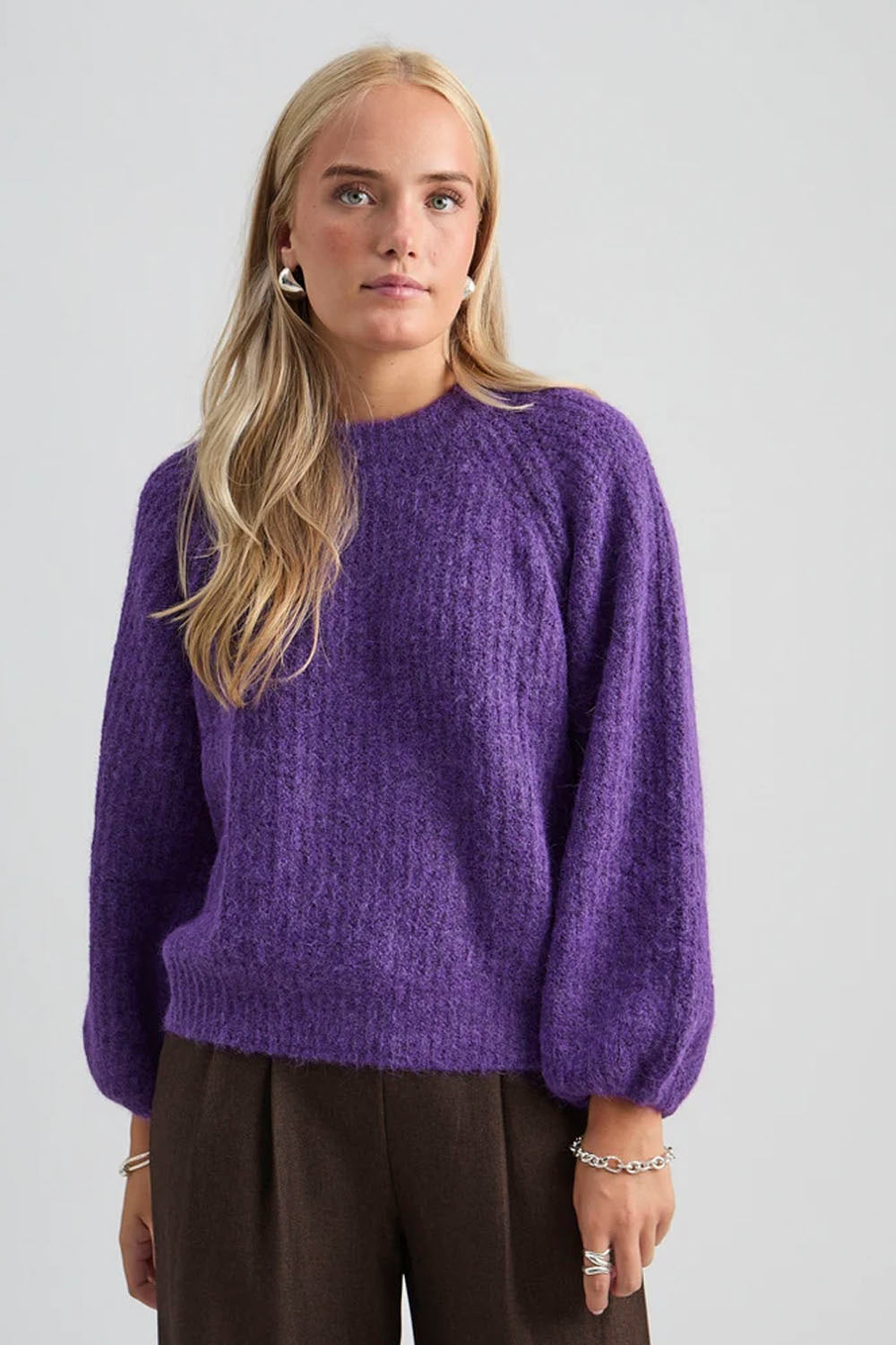 A woman wearing a textured, fuzzy purple knit sweater with balloon sleeves, paired with dark brown trousers, against a light gray studio background.