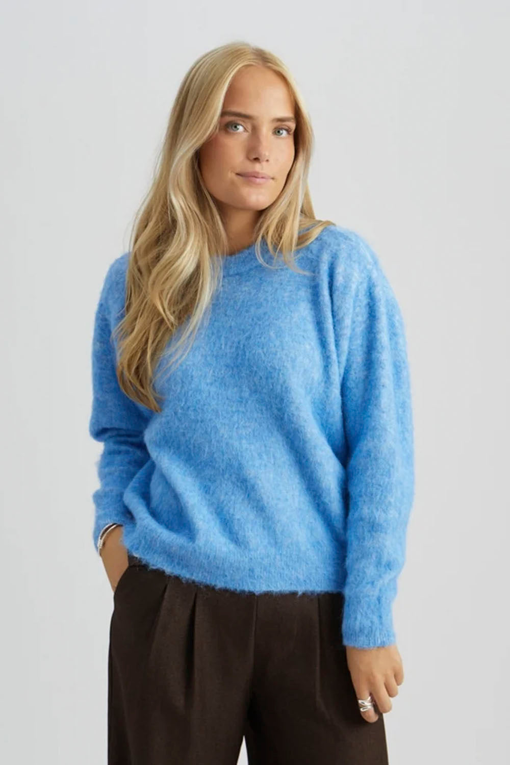 A woman with long blonde hair wearing a fuzzy light blue crewneck sweater and dark brown trousers, standing against a light gray studio background.