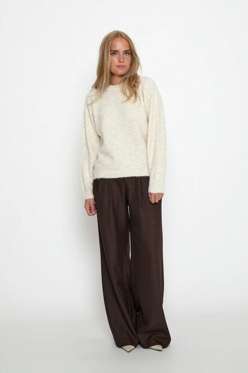 A woman stands against a plain white studio background wearing a cream knit sweater and dark brown wide-leg trousers.