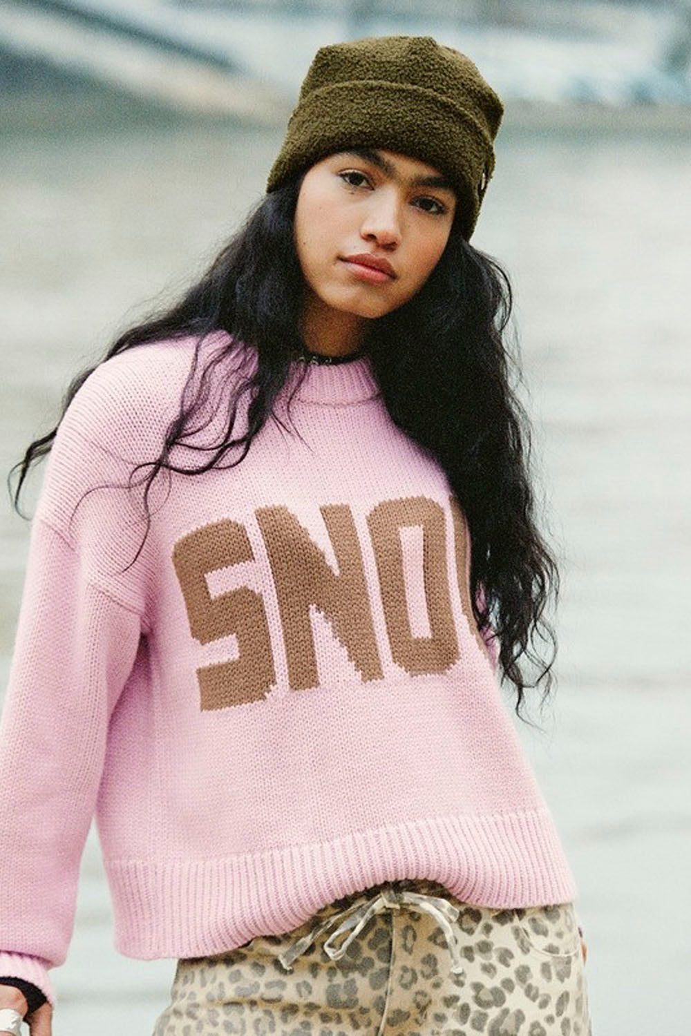 A woman wearing a pink knit sweater with 'SNO' in brown lettering, a brown beanie, and leopard-print pants, photographed outdoors against a blurred gray background.