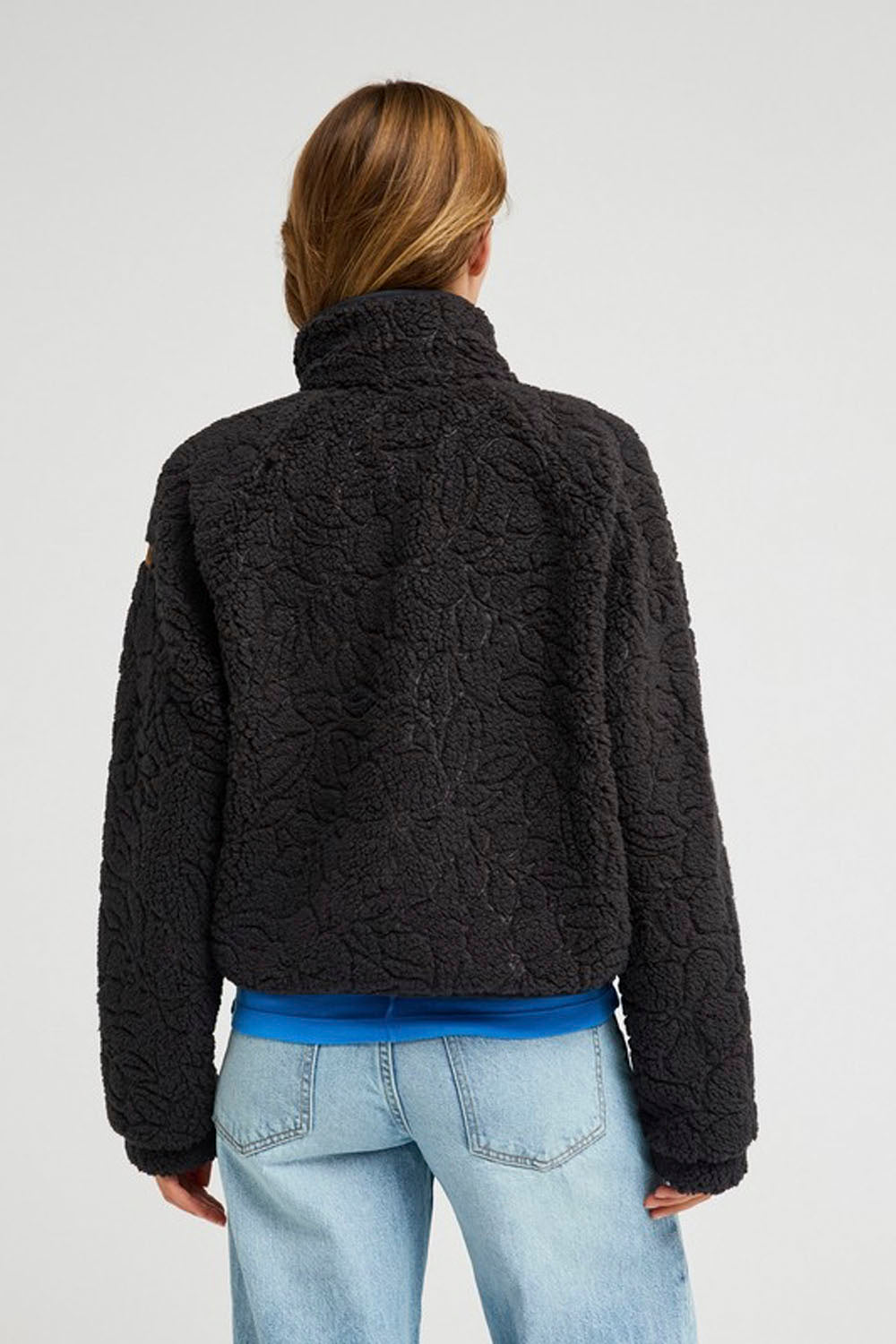 A woman viewed from the back wearing a black textured boucle fleece jacket, a bright blue t-shirt, and light wash blue jeans against a light gray studio background.
