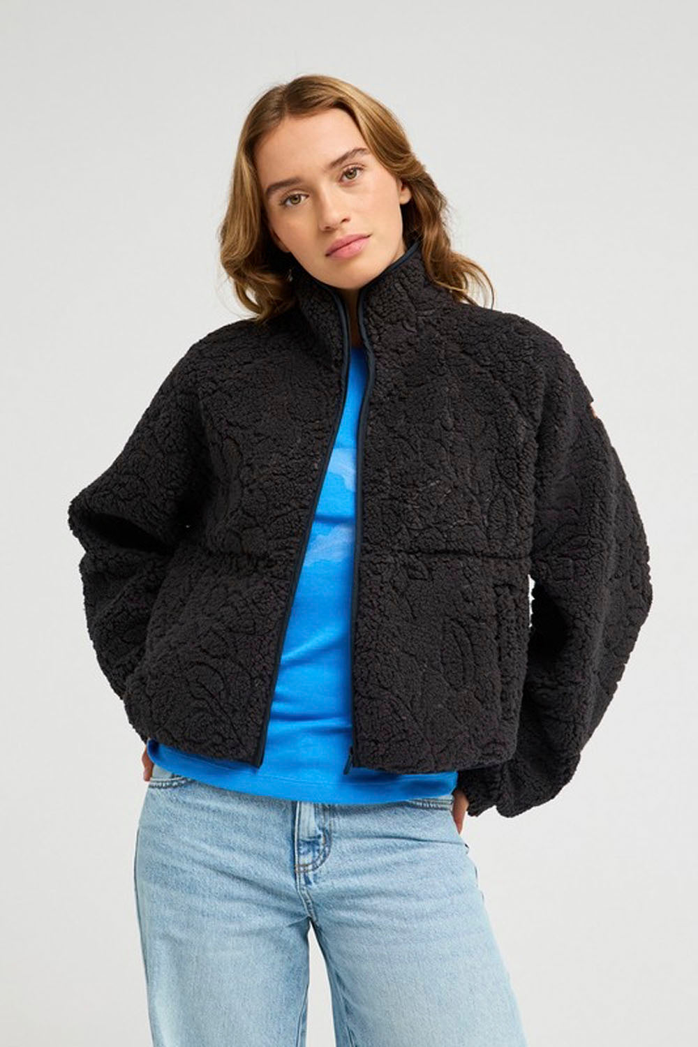 A woman wearing a black textured teddy fleece jacket over a bright blue top and light wash jeans, photographed against a light gray studio background.