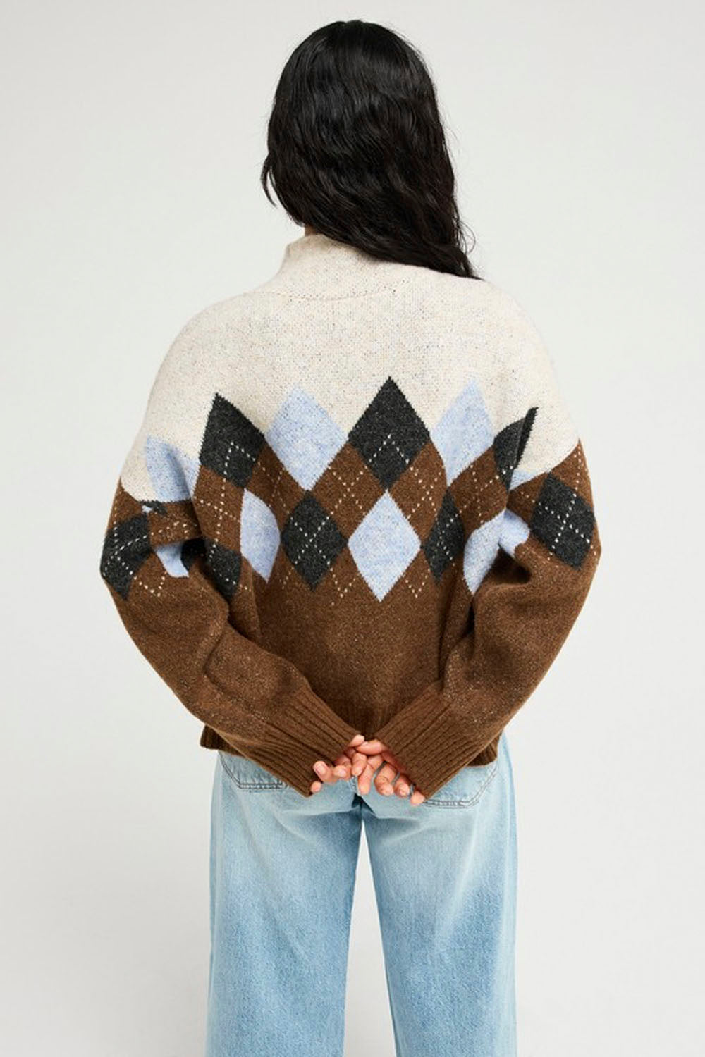 A person viewed from the back wearing a brown knit sweater with a cream upper section and an argyle diamond pattern in brown, black, light blue, and white across the shoulders and upper back, paired with light blue jeans against a plain light gray studio background.