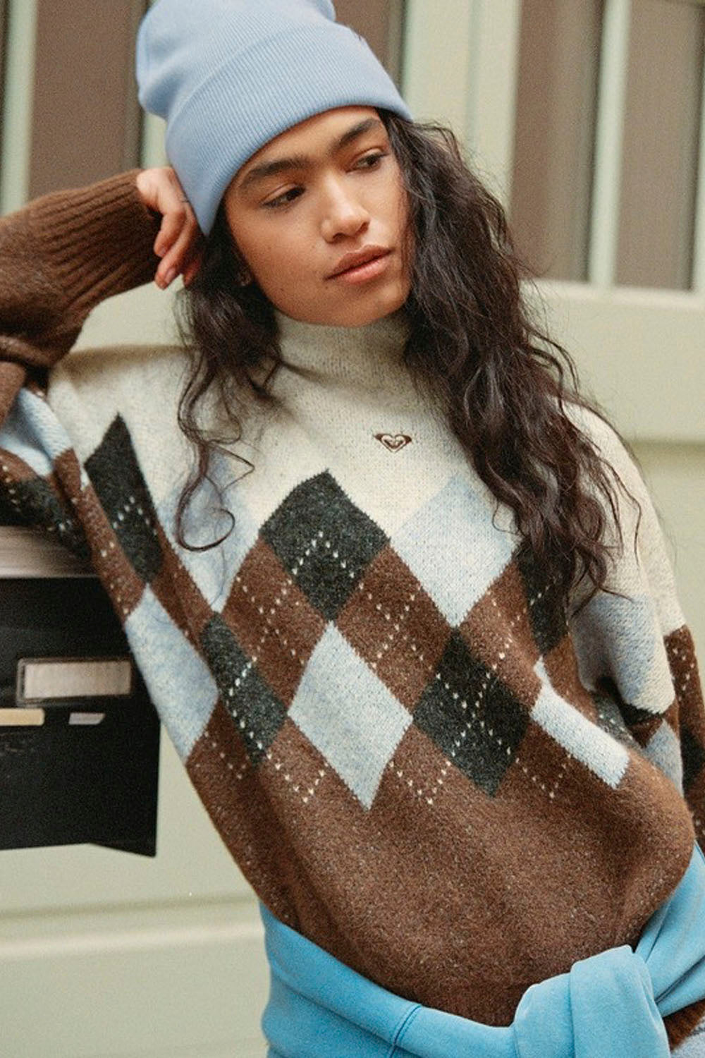 A woman wearing a brown, beige, and black argyle knit sweater with a light blue beanie and a matching light blue tie belt, posed outdoors next to a black mailbox.