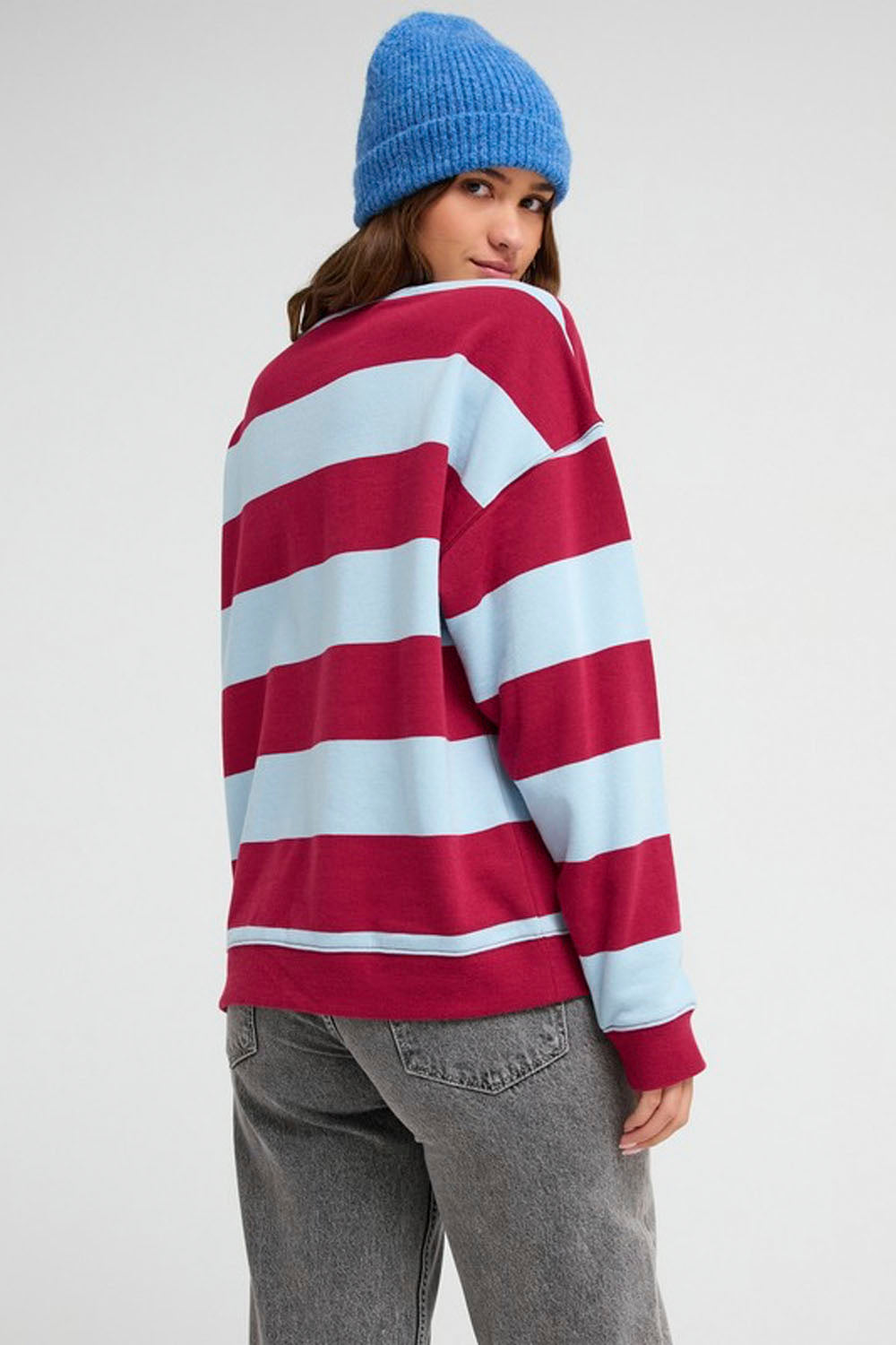 A woman wearing a red and light blue horizontally striped long-sleeve sweatshirt, gray jeans, and a blue knit beanie, photographed against a plain light gray studio background.