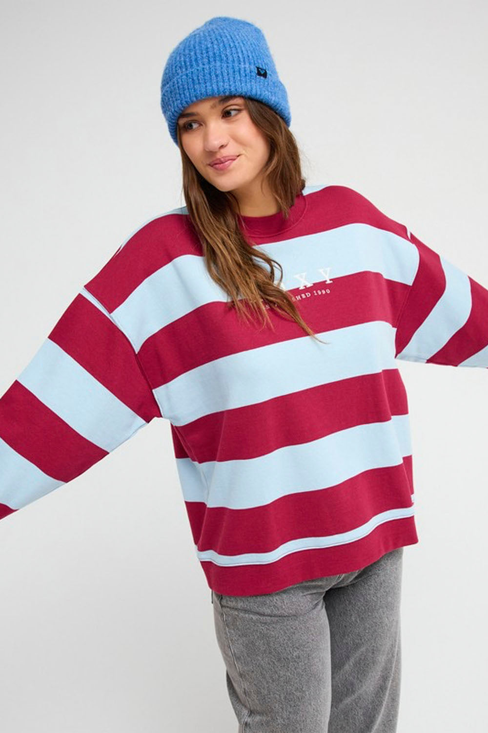 A woman wearing a red and light blue horizontally striped sweatshirt, gray sweatpants, and a blue knit beanie against a light gray studio background.