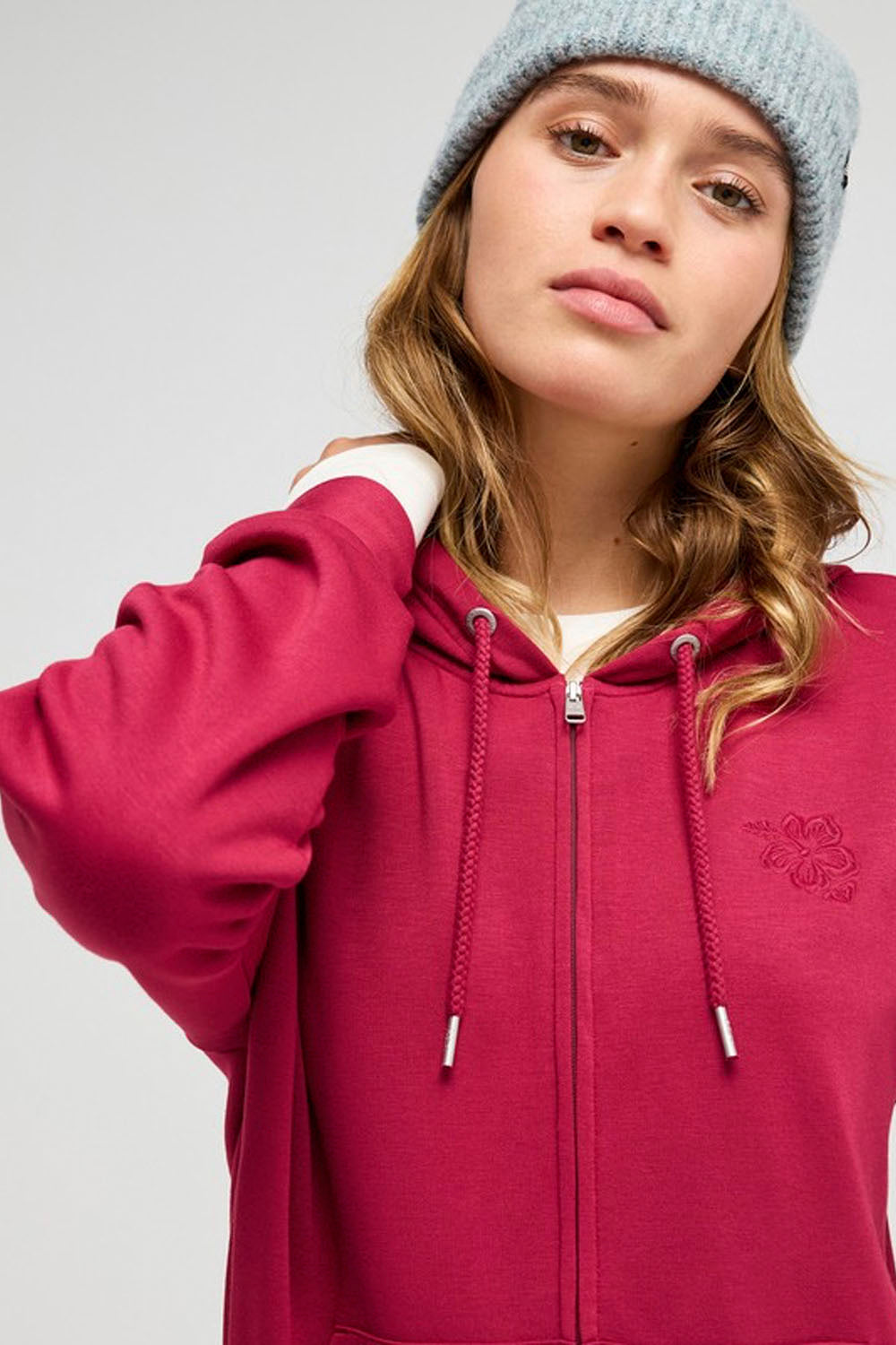 A woman wearing a bright pink zip-up hoodie with a small embroidered rose on the left chest and a light blue knit beanie, photographed against a neutral light gray background.