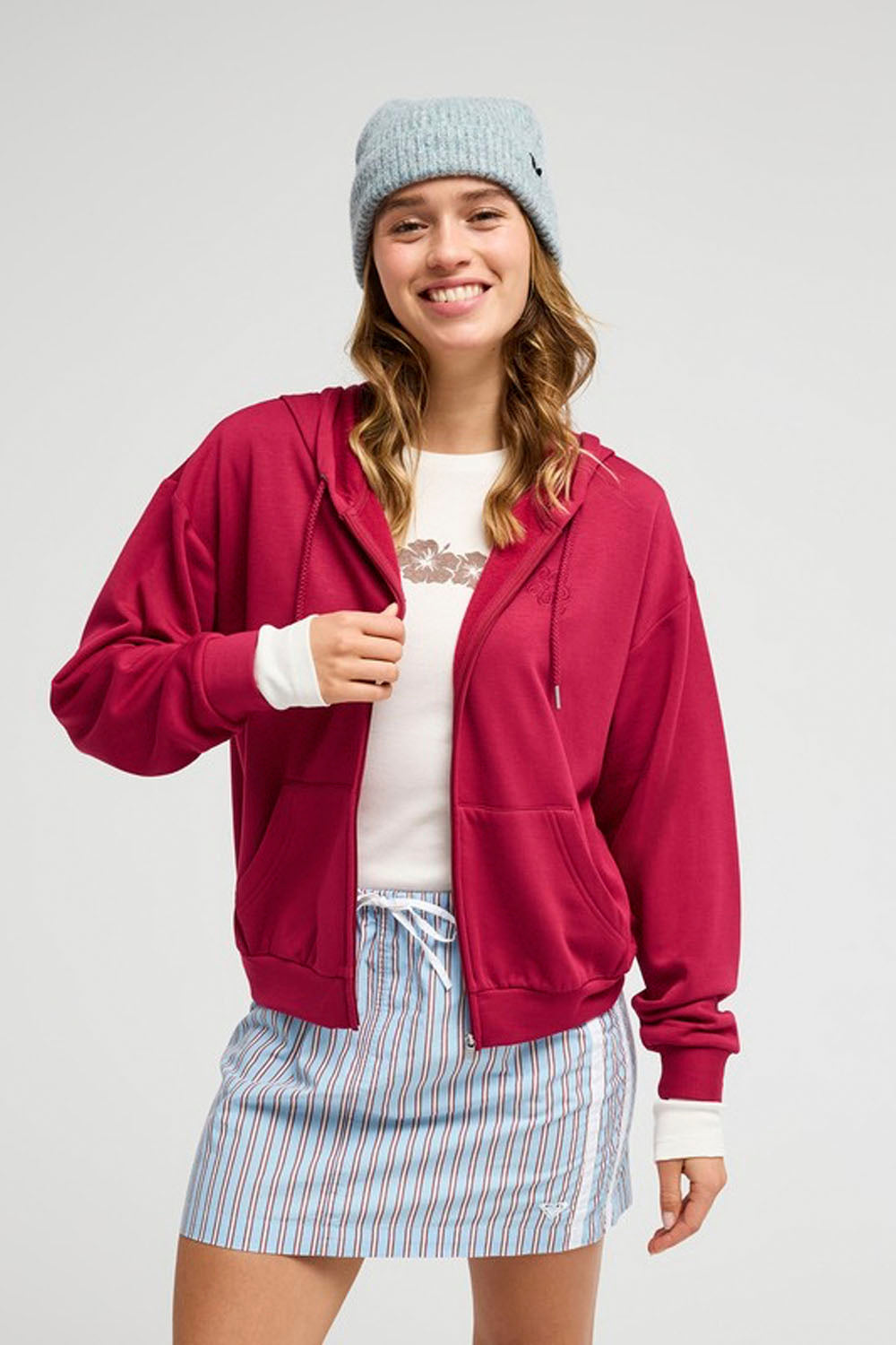 A woman wearing a bright magenta zip-up hoodie, a light blue knit beanie, a white graphic t-shirt, and a blue and white striped mini skirt, posing against a light gray studio background.