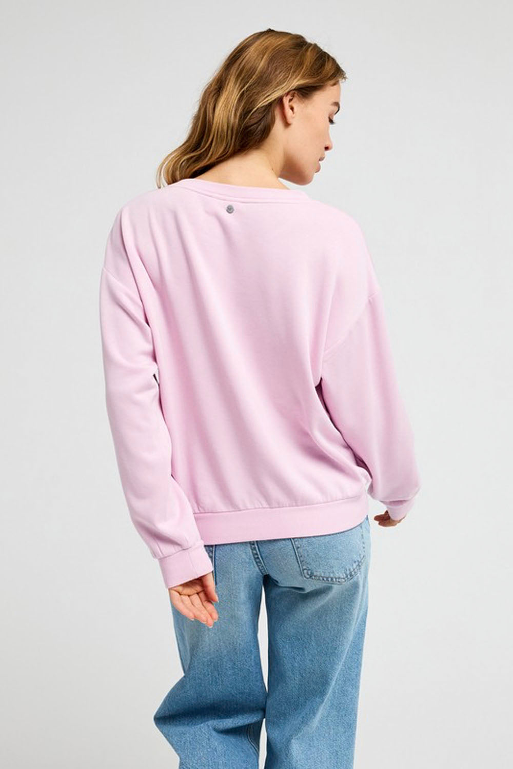 A woman shown from the back wearing a light pink crewneck sweatshirt and blue jeans against a light gray studio background.