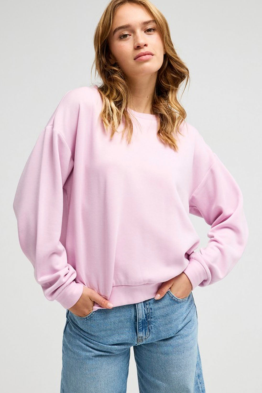 A woman wearing a light pink crewneck sweatshirt and blue jeans, standing against a light gray studio background.