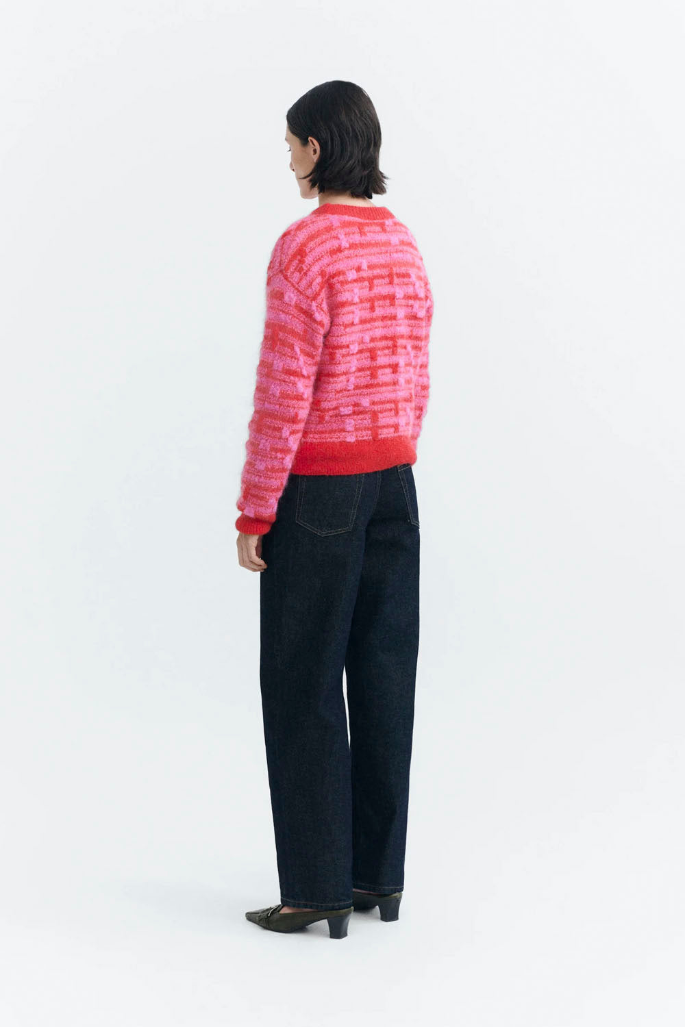 A model shown from the back wearing a pink and red striped knit sweater, dark wide-leg jeans, and black heels, photographed against a clean white studio background.