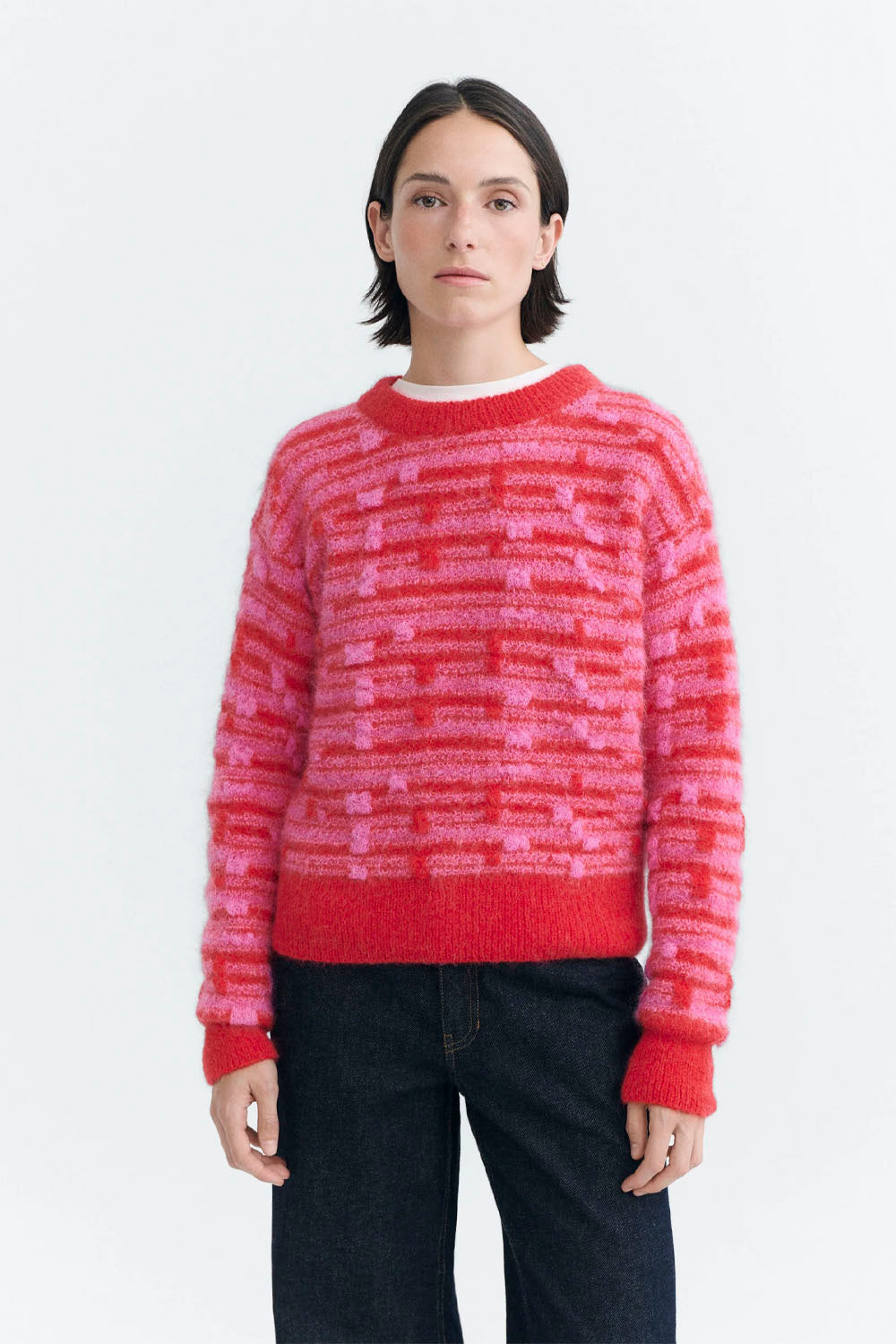 A model wearing a fuzzy knit crewneck sweater with horizontal stripes of red, pink, and orange, paired with dark blue jeans, against a plain white background.