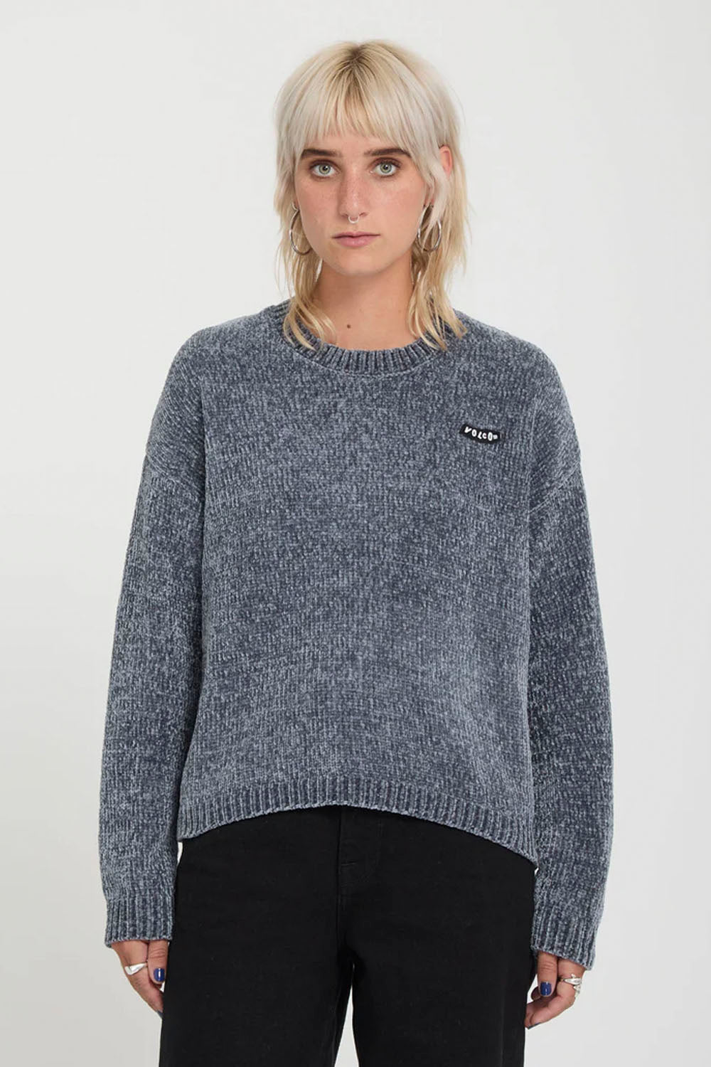 A model with short blonde hair wearing a gray marled knit sweater with a small black and white logo patch on the left chest, paired with black pants, against a plain light gray background.
