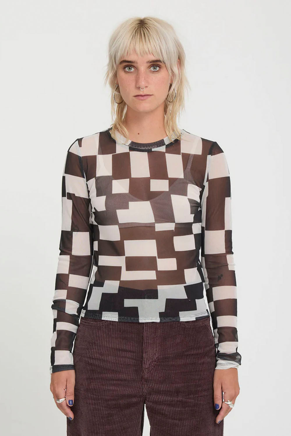A woman modeling a sheer long-sleeve top with a brown, beige, and black geometric block pattern, paired with dark brown corduroy pants, against a plain white studio background.
