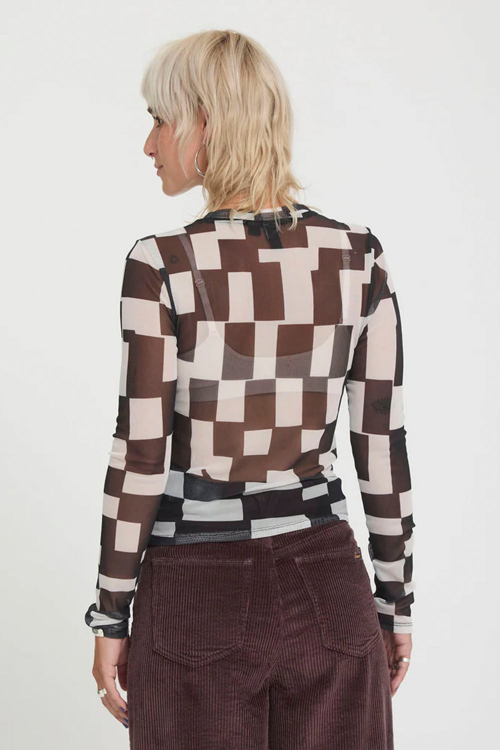 A woman with short blonde hair is shown from the back wearing a sheer long-sleeve top with a brown, black, and beige geometric block pattern, paired with dark brown corduroy pants, against a plain white background.