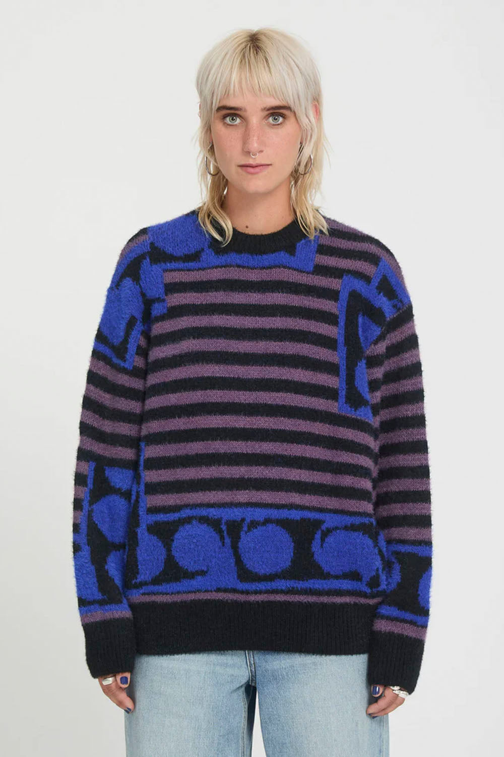 A model with blonde hair wearing a purple and black striped knit sweater with blue geometric patterns, paired with light blue jeans, against a plain white background.