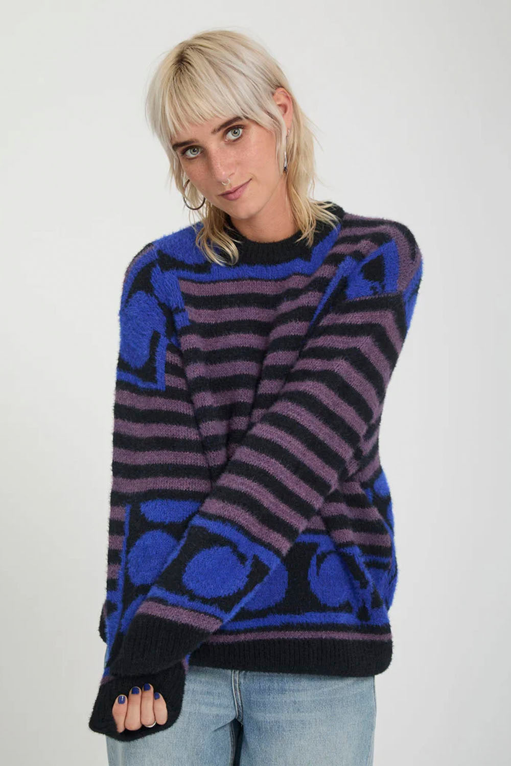 A woman wearing a fuzzy knit sweater with horizontal stripes and abstract geometric patterns in purple, blue, and black, paired with light-wash jeans against a plain light gray background.