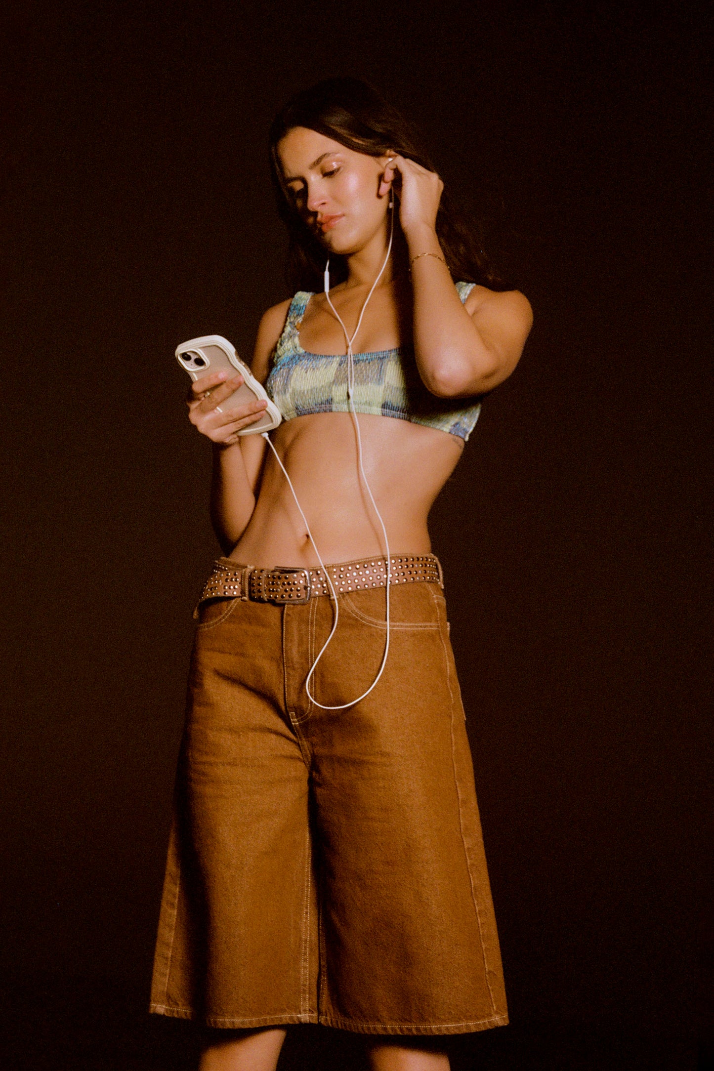 Woman wearing a plaid top and brown pants, holding a phone and earphones against a dark background