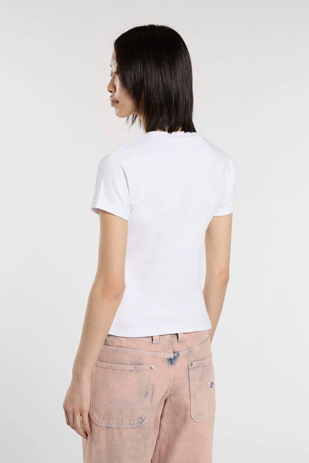 A woman with short dark hair is shown from the back wearing a plain white short-sleeve t-shirt and light pink jeans against a light gray studio background.