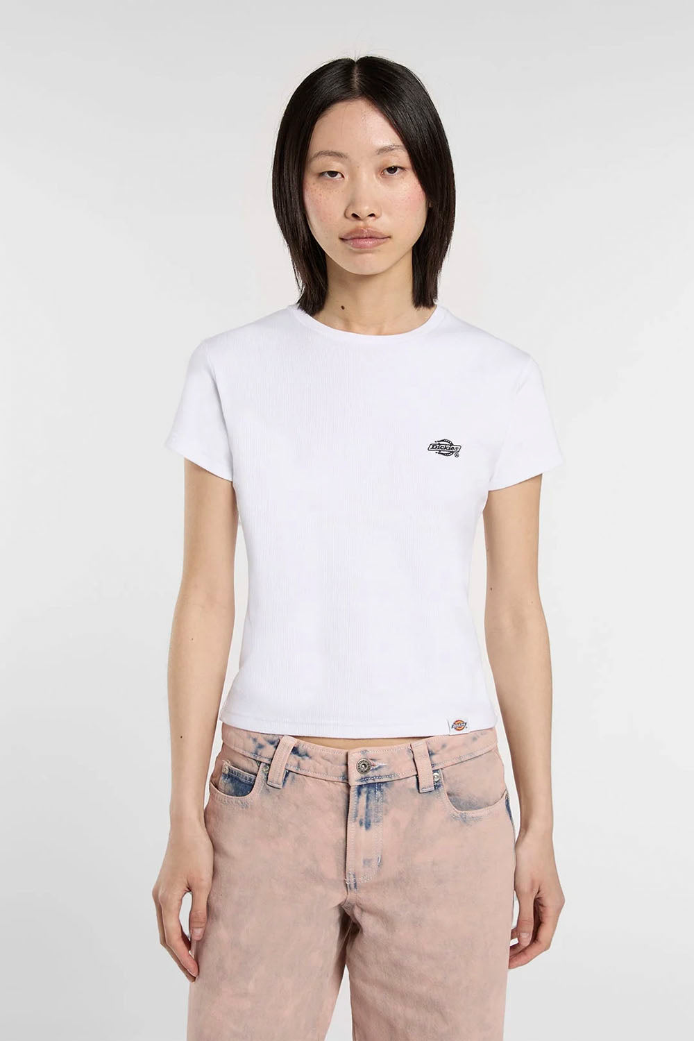 A model wearing a white short-sleeve crewneck t-shirt with a small fish logo on the left chest, paired with light pink jeans, against a plain light gray background.