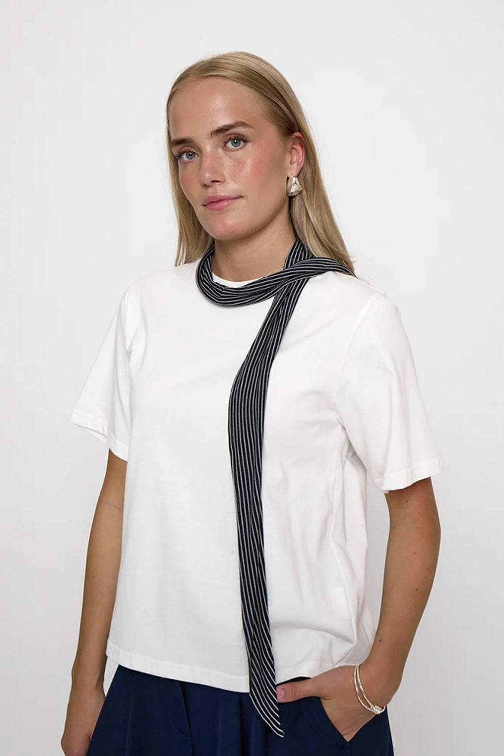 A woman modeling a white short-sleeve t-shirt with a black and white vertically striped scarf wrapped around her neck, set against a plain white studio background.