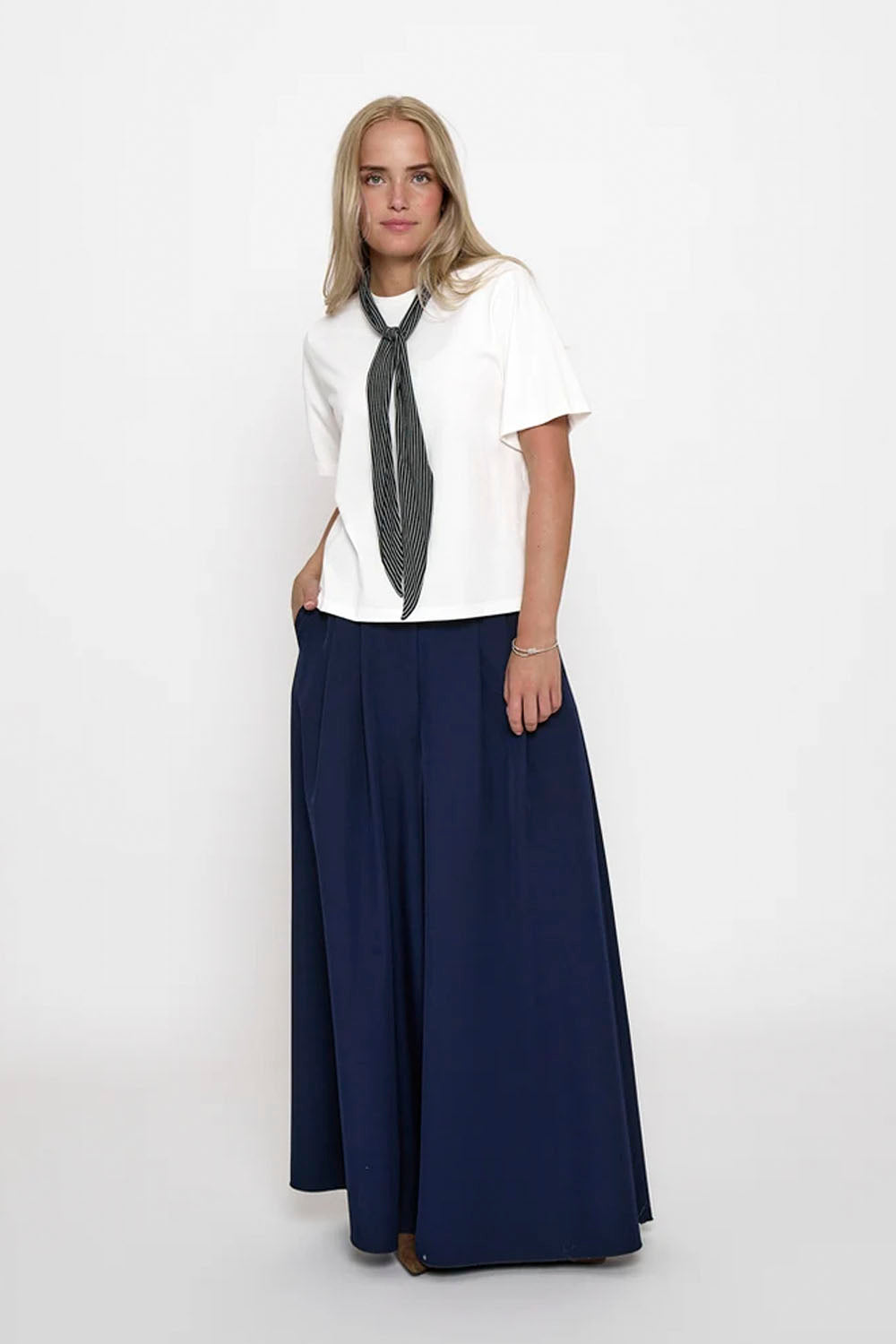 A woman modeling a white short-sleeve top with a dark gray patterned neck scarf and high-waisted navy blue wide-leg trousers, standing against a plain white studio background.