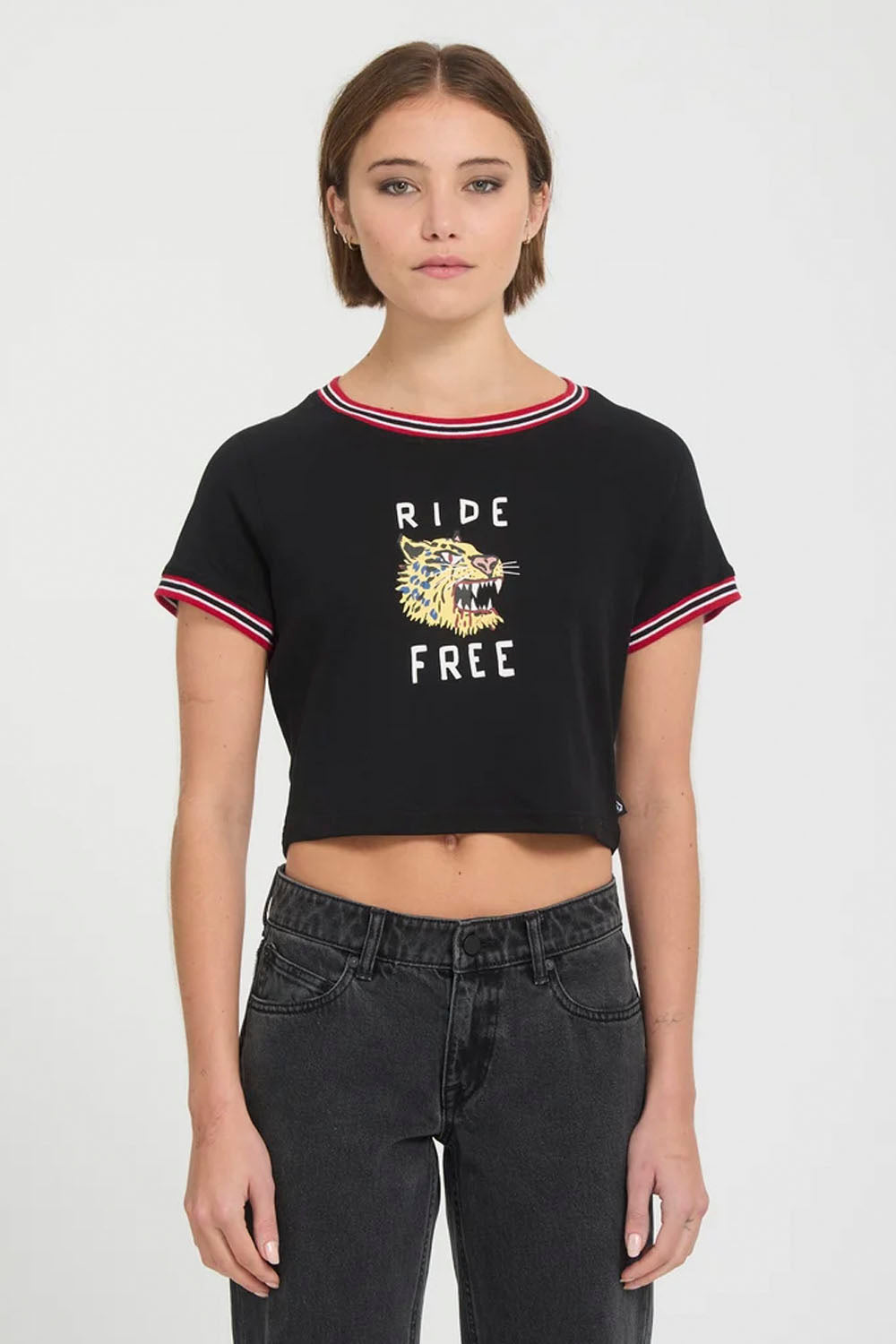 A model wearing a black short-sleeve crop top with red and white striped ruffle trim, featuring a graphic of a tiger and the text 'RIDE FREE'. She is also wearing dark gray jeans against a plain light gray background.