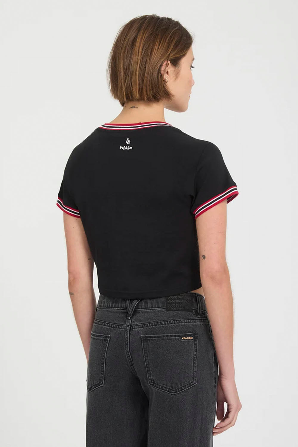 A woman shown from the back wearing a black cropped t-shirt with red and white striped trim on the collar and sleeve cuffs, paired with dark gray jeans against a plain white studio background.