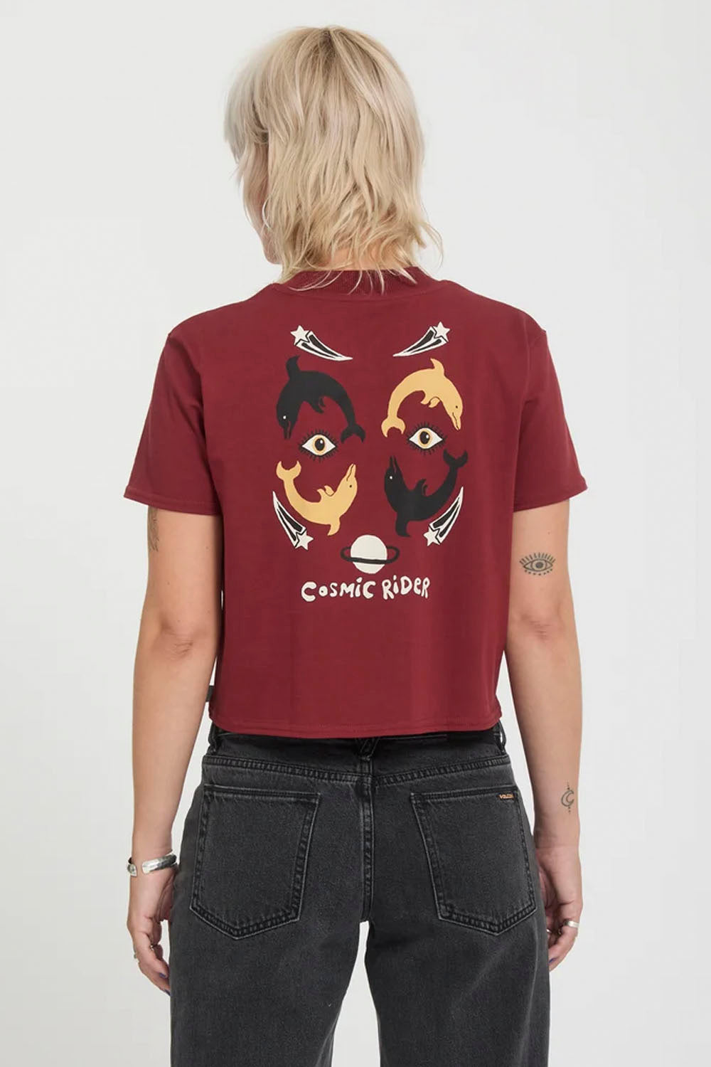 A model from behind wearing a cropped maroon t-shirt with a graphic of stylized faces and the text 'COSMIC RIDE' on the back, paired with dark gray jeans against a white background.