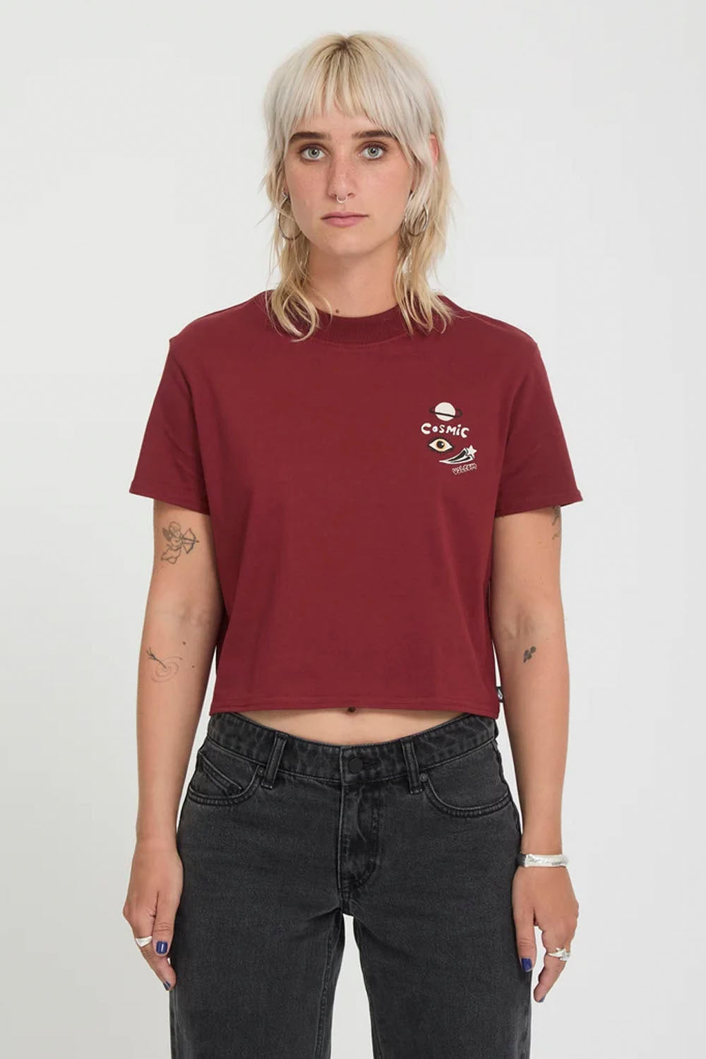 A model with short blonde hair wearing a cropped maroon t-shirt with a small white graphic on the left chest, paired with dark gray jeans, against a plain white background.
