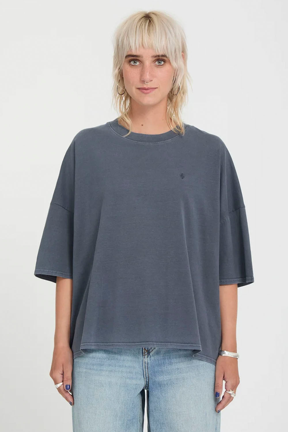 A model with short blonde hair wearing a dark slate blue-gray oversized t-shirt and light-wash blue jeans, standing against a plain white studio background.