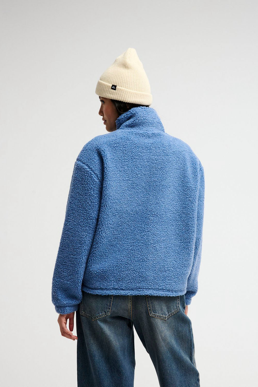 A person shown from the back wearing a textured blue fleece pullover, a cream knit beanie, and blue jeans against a plain light gray studio background.