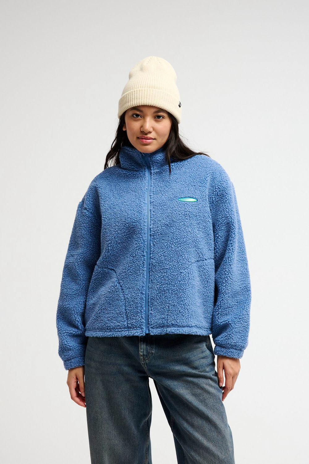 A woman wearing a blue sherpa fleece zip-up jacket, a cream knit beanie, and dark gray jeans, standing against a plain light gray studio background.