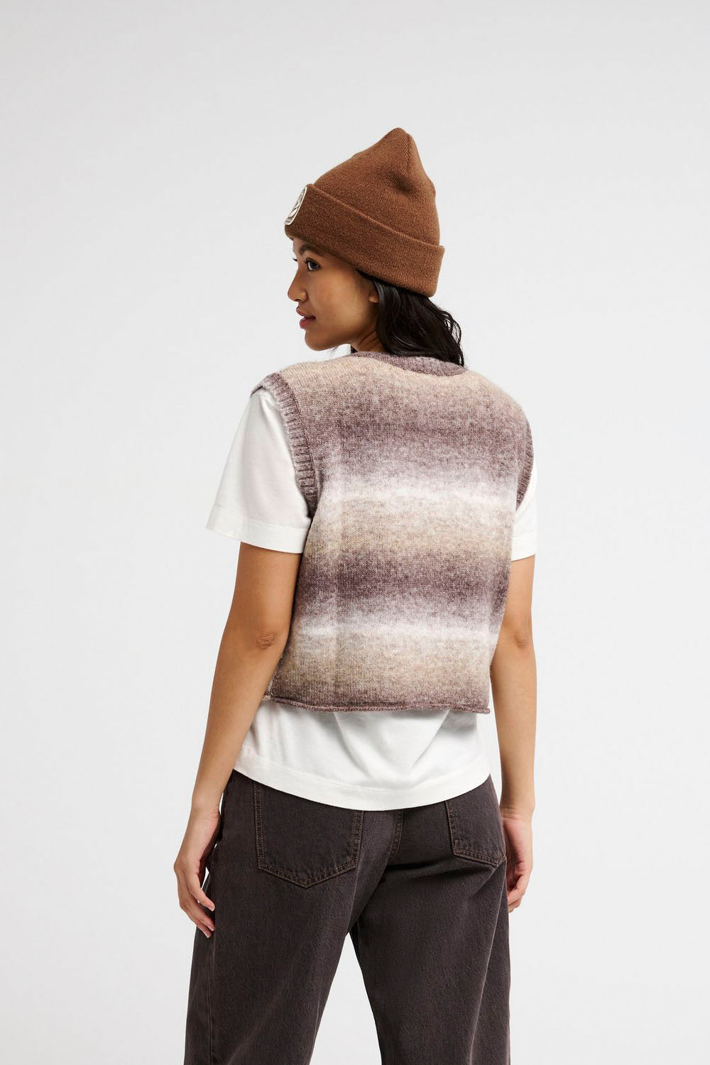 A model shown from the back wearing a brown knit beanie, a white t-shirt, and a brown and beige striped knit sweater vest over dark brown pants, against a plain white studio background.