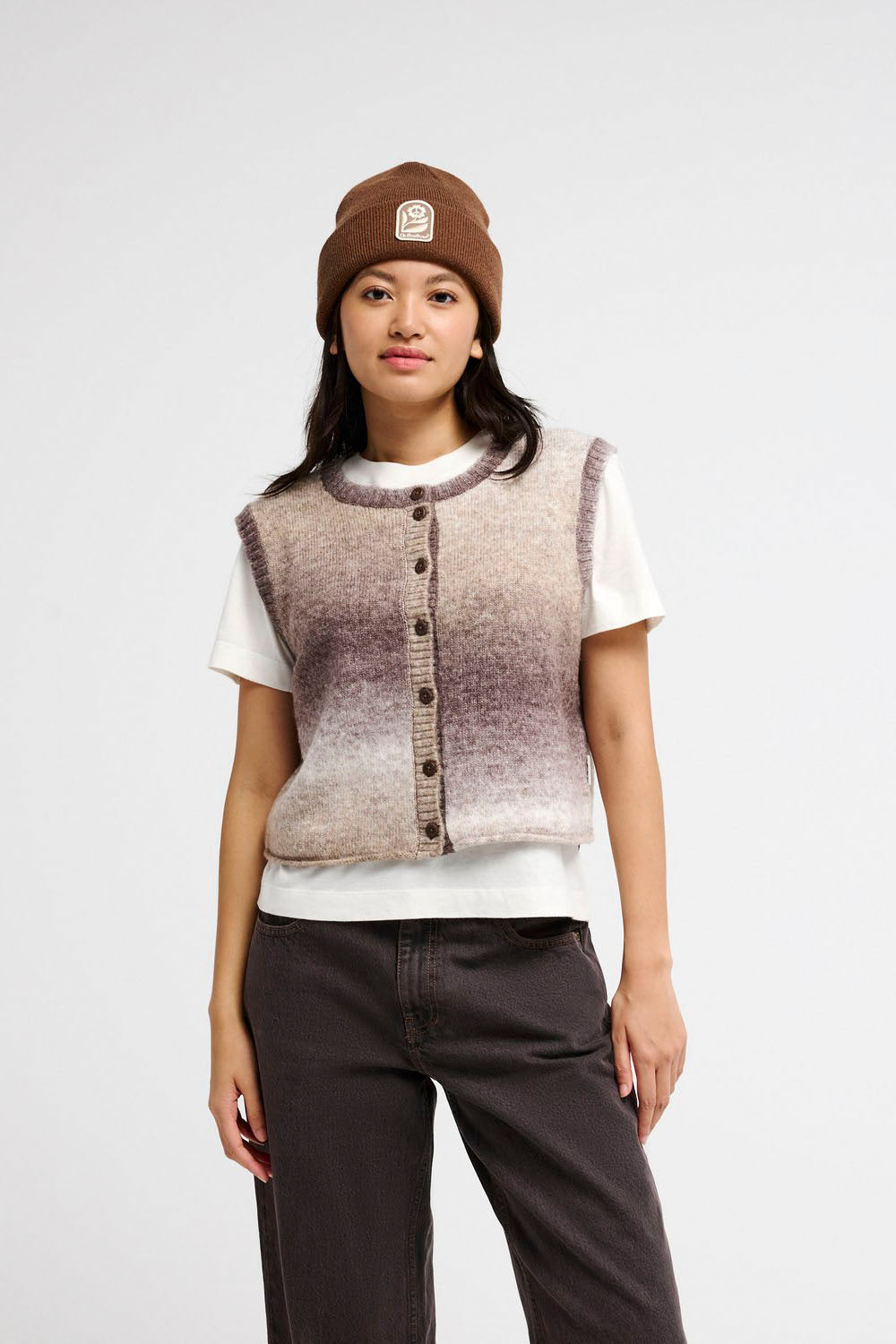A model stands against a light gray studio background wearing a brown knit beanie, a sleeveless fuzzy vest over a white t-shirt, dark wide-leg cropped pants, and black sneakers.