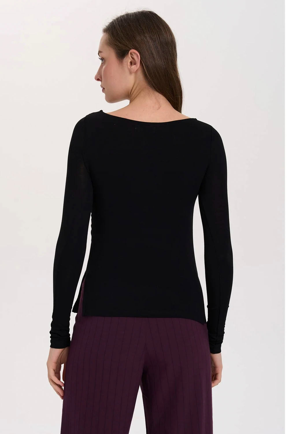 A woman modeling a black long-sleeve boat-neck top with gathered detailing at the waist, paired with dark purple pinstriped pants, against a plain white studio background.