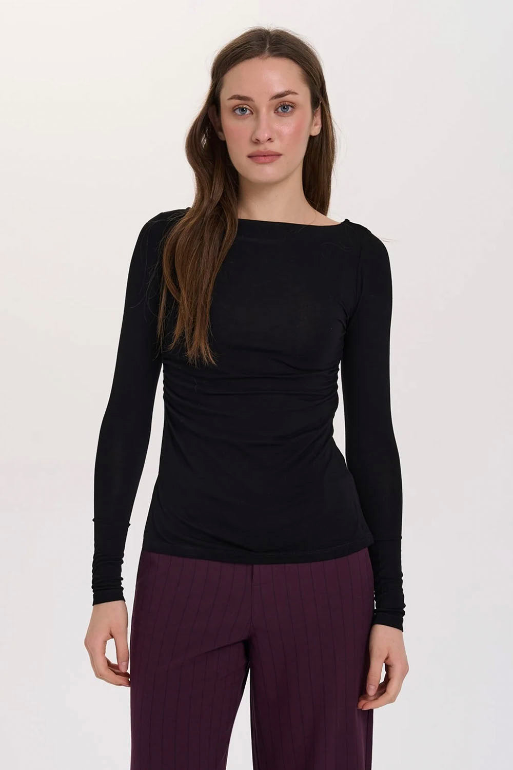 A woman modeling a black long-sleeve boat-neck top with gathered detailing at the waist, paired with dark purple pinstriped pants, against a plain white studio background.